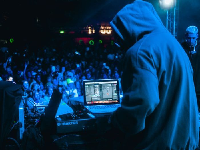 DJ in hoodie performs on stage at night music festival with energetic crowd and colorful lights.