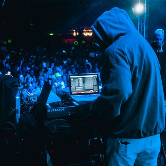 DJ in hoodie performs on stage at night music festival with energetic crowd and colorful lights.
